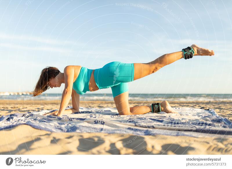 A woman performs leg pull front movement whit extra weight in the ankles while practicing Pilates on the beach exercise pilates yoga mediterranean spain
