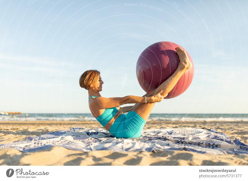 A woman practices the double-leg stretch movement on the beach while holding a Pilates ball abdominal legs exercise pilates yoga mediterranean spain