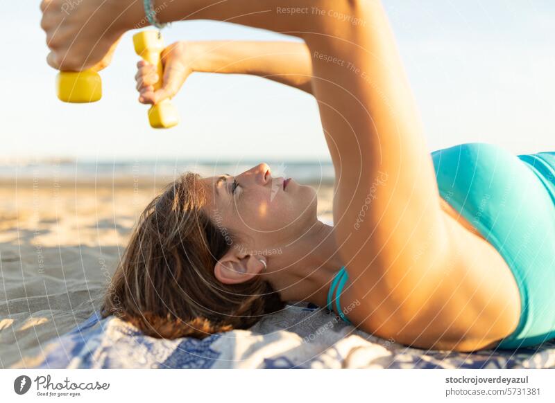 A woman performs stretching and relaxation exercises with light 1kg dumbell weights, lying on the beach dumbbell pilates yoga mediterranean spain