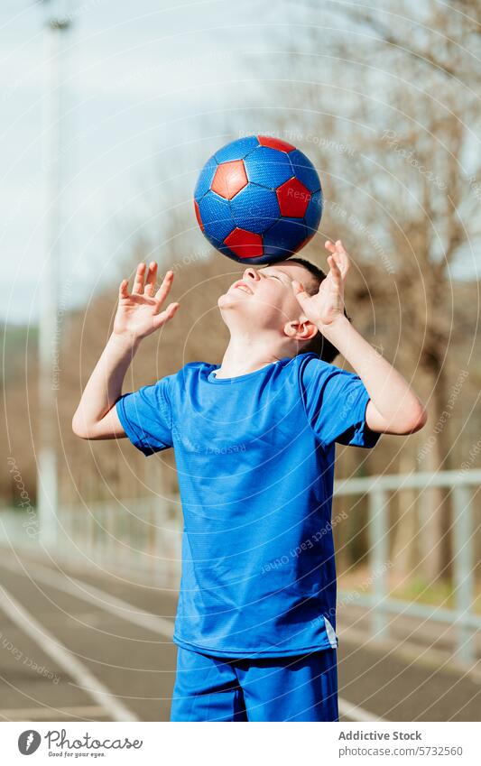 Young boy balancing soccer ball on head outdoors sports uniform blue red determined expression clear sky background young child kid play skill practice training