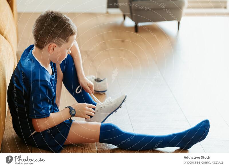 Young boy in soccer gear tying shoelaces indoors uniform sport sitting floor wooden blue white child sportswear determination focus youth soccer player training