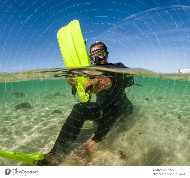 A snorkeler in a black suit with a vibrant yellow fin peers through a diving mask, half-submerged in sunlit, clear sea water woman bright fin surface adventure