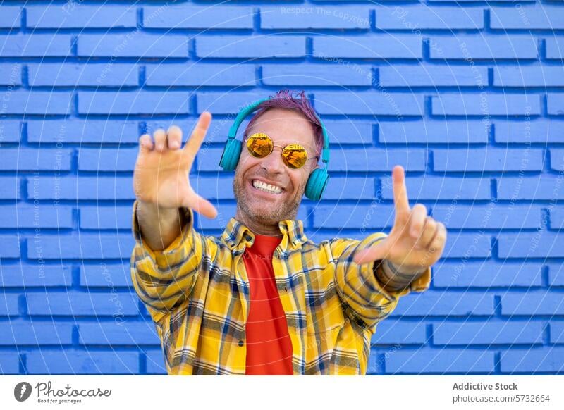 A playful man in colorful headphones throws up a rock sign, enjoying music with a blue brick wall as his vibrant stage groove beat yellow checkered shirt