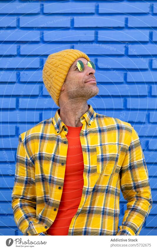 Cheerful man in yellow beanie and sunglasses looks up, basking in the sunlight against a textured blue brick wall backdrop sunshine cheerful portait happy joy