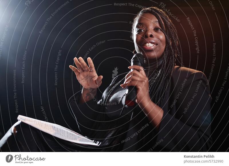 Waist up portrait of African American woman holding microphone and wearing smart black suit while speaking on stage, copy space Black woman speaker politics
