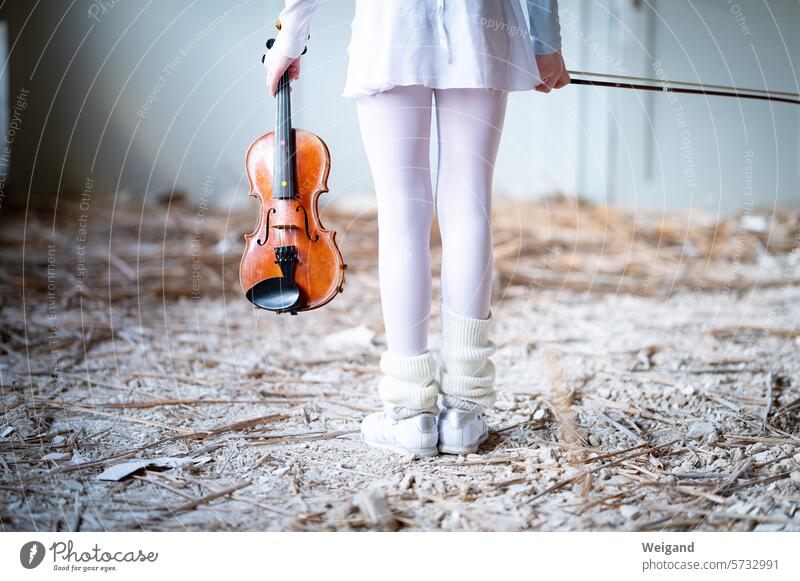 Little girl dressed all in white like a ballerina, standing in the middle of a construction site with her back to the photographer and holding a wooden violin in her hand to play music in the creative chaos