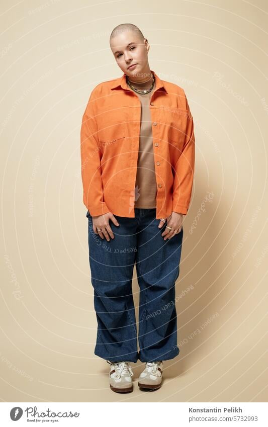 Vertical full length portrait of bald young woman wearing bright orange jacket and baggy jeans posing in studio looking at camera female girl gen Z hair loss