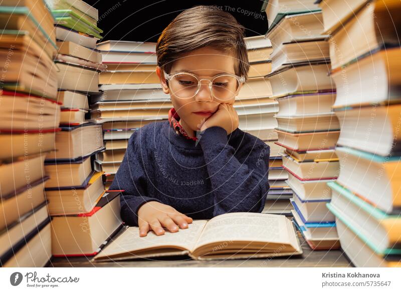Cute little pupil boy in glasses reading interesting book in library between stacks of books literature. Education concept, prep or elementary school education