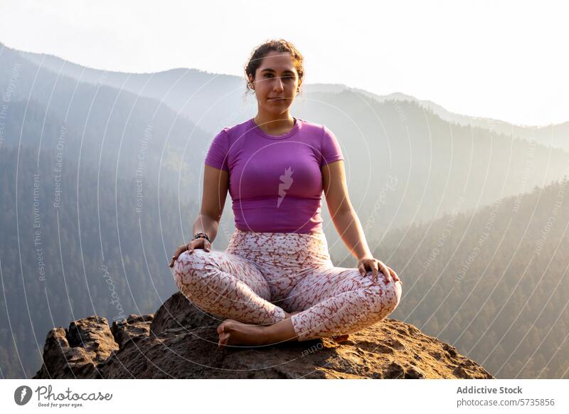 A woman in a yoga pose sits tranquilly atop a mountain, as the setting sun casts a warm glow over the forested landscape meditation sunset tranquility nature