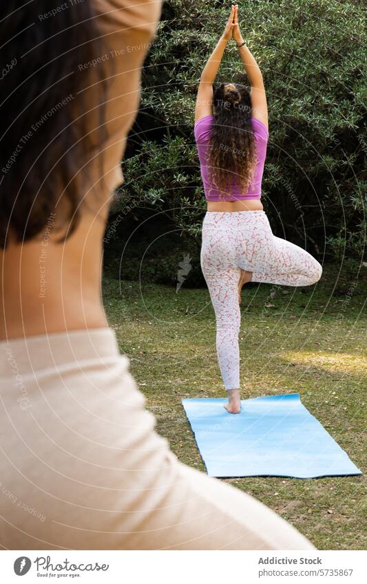 Anonymous woman practices the tree pose in yoga, finding balance on her mat in a peaceful natural environment as another observes observer nature outdoor health
