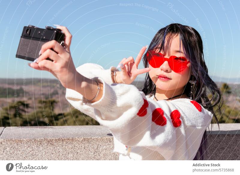 Young chinese girl taking selfie with vintage camera outdoors gen-z heart-shaped sunglasses sweater hearts young retro clear sky fashion style trendy leisure