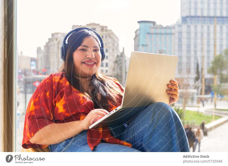 Smiling woman with laptop and headphones in urban setting city music cheerful environment sunny building smile technology online leisure casual wireless