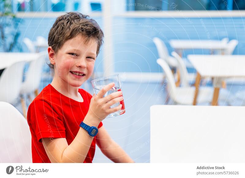 Smiling boy drinking water in a bright cafe setting child glass smile happy seated indoor red shirt refreshment hydration health wellbeing joy table chair blur