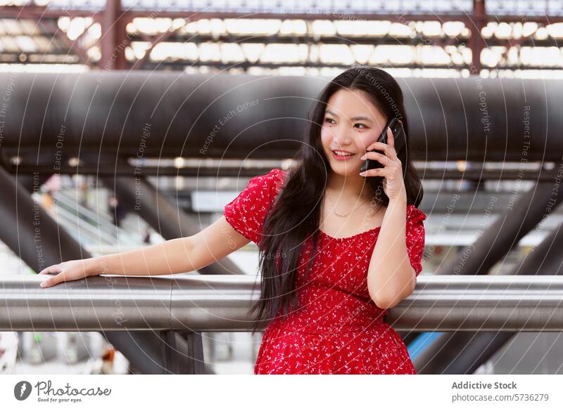 Smiling young woman talking on phone in urban setting mobile phone conversation smiling red dress railing metal blurred background city infrastructure leaning