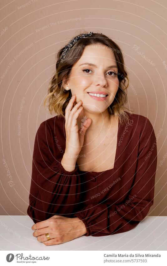 Young woman smiling, with brown hair and brown blouse on isolated beige background. Using hair accessories. 30-35s amazed beautiful beauty salon