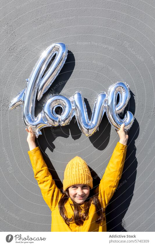 Young girl holding silver love balloon against a wall yellow hat coat happy concrete gray joyful moment celebration young lifted shiny metallic decoration