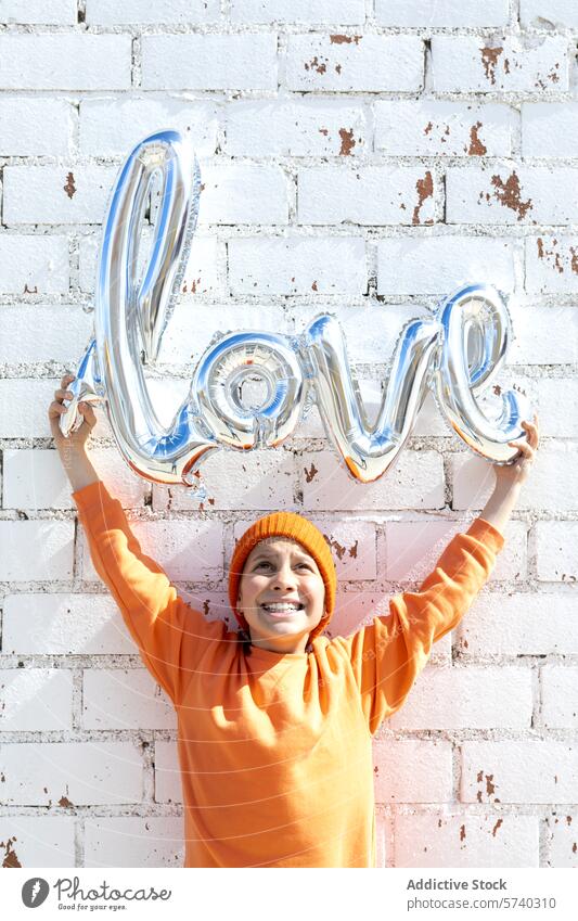 Woman holding a love balloon against a brick wall woman portrait white silver joyful orange hoodie hat happiness celebration smiling cheerful outdoors day