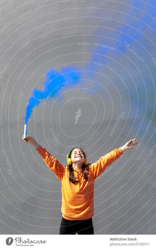 Joyful woman with colorful smoke flare in urban setting joy happiness celebration headphones music young outdoor blue smile raise arms enjoyment orange hoodie