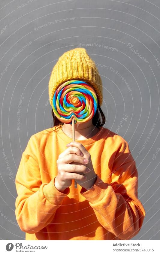 Person with colorful lollipop covering their face person candy sweet treat hidden face obscured disguise playful vibrant yellow beanie orange sweatshirt
