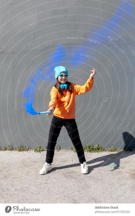 Youthful spirit in casual urban style with vibrant colors woman young cheerful dance gray wall orange sweatshirt blue beanie sunglasses blue smoke smoke flare