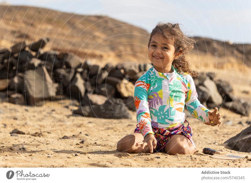 A happy toddler girl in a colorful swimsuit plays with sand on a beach, with rocks and hills in the background outdoor leisure child sea coast sunny playful