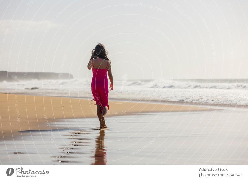 Anonymous young girl in a pink dress walks alone along the shore, leaving footprints in the wet sand as the waves gently break beach sunset solitary sea ocean