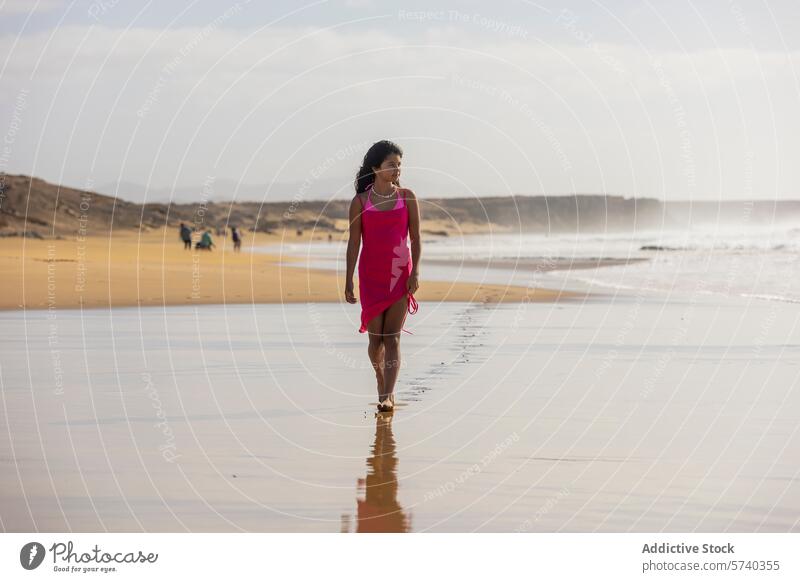 A young girl in a pink dress walks alone along the shore, leaving footprints in the wet sand as the waves gently break beach sunset solitary sea ocean coast