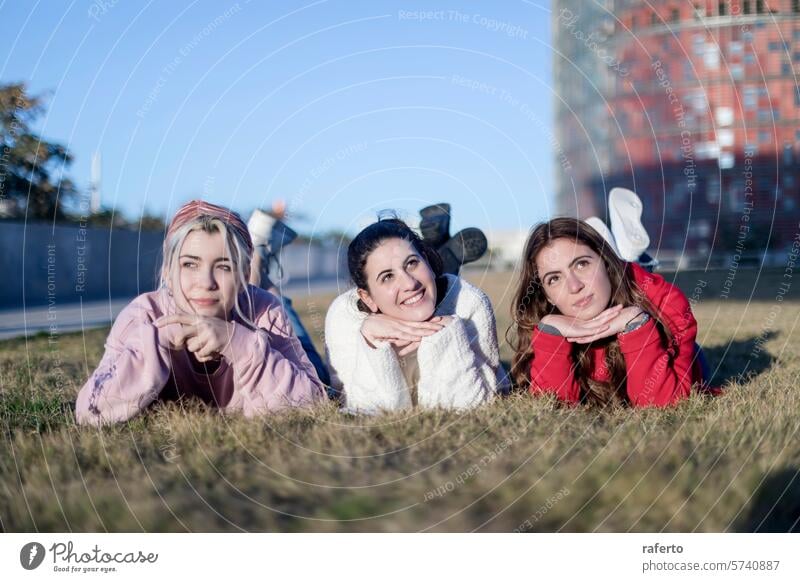 Three friends posing playfully on the grass young women clear sky urban background lying down stomachs hands chins casual outdoors leisure day happy cheerful