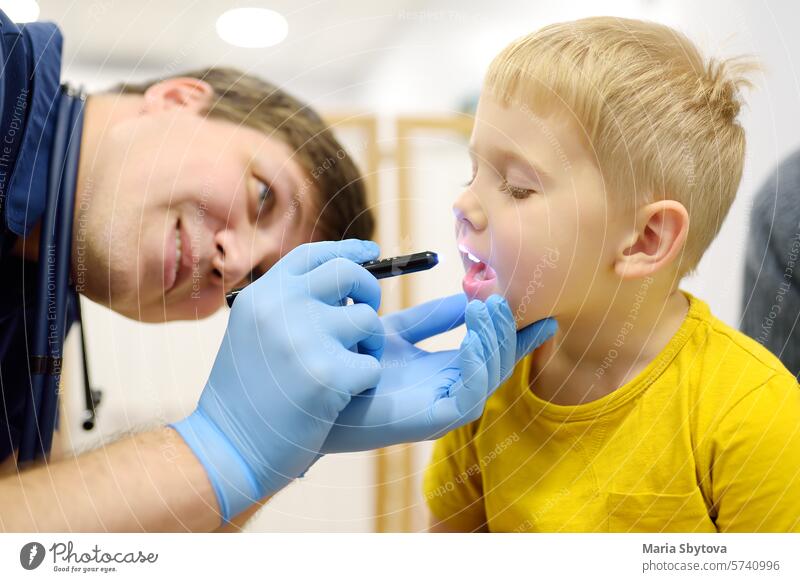 A cute toddler boy with his mother are at an appointment with a pediatrician. The kids ENT doctor examines the throat of a little patient using a flashlight.