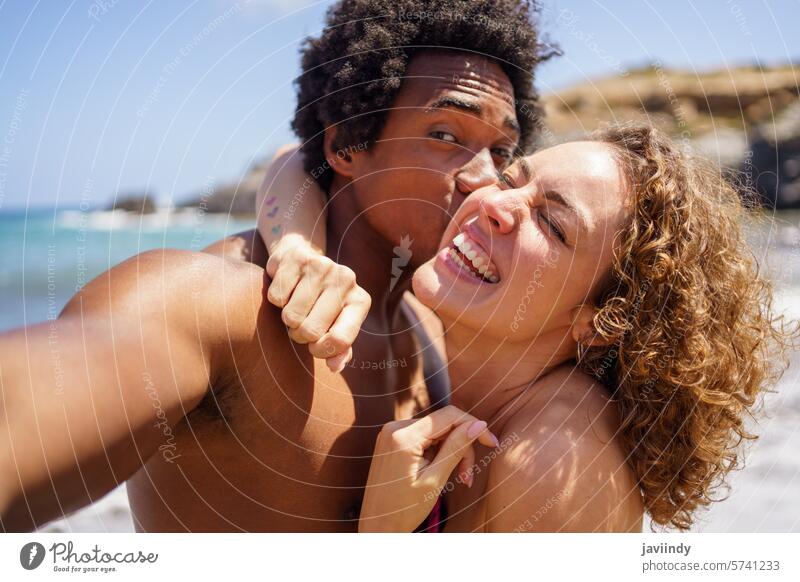 Black man kissing cheek of happy girlfriend while taking selfie couple take photo hug holiday delight sunlight beach resort smile multiethnic african american