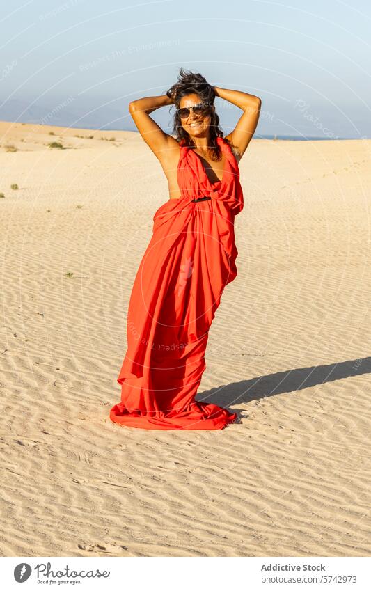Woman in flowing red dress enjoys sunshine in desert woman dunes vibrant stand hands hips sandy clear sky smiling sunglasses fashion style elegant flowy