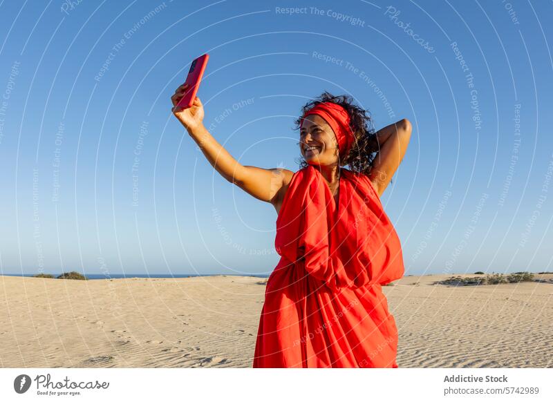 Joyful woman taking selfie in desert with phone red dress dune mobile phone smile joy happiness blue sky gadget device summer outdoor sunny technology