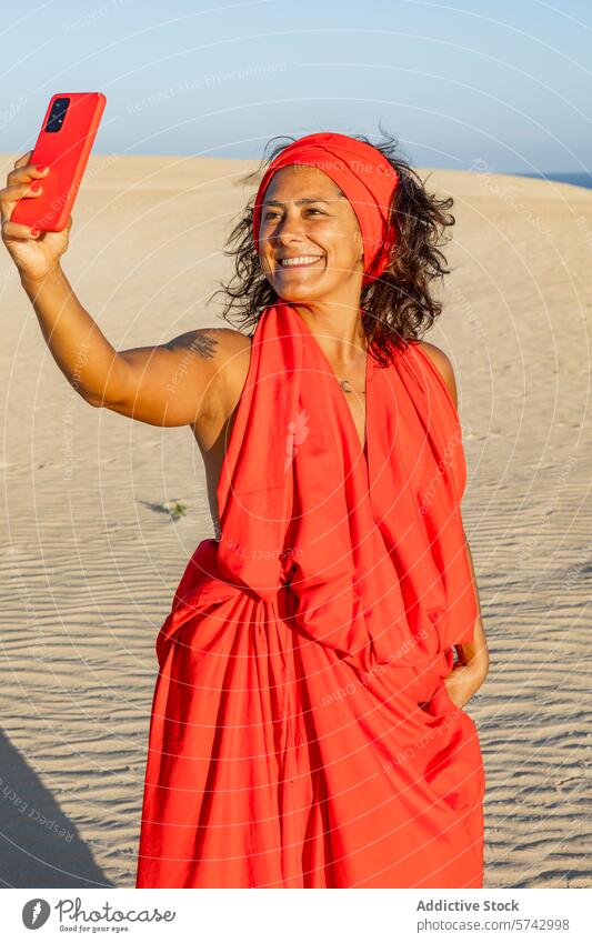 Woman in Red Dress Taking Selfie in Desert woman red dress desert dune selfie mobile phone headscarf smile joyous sandy screen looking at screen device gadget