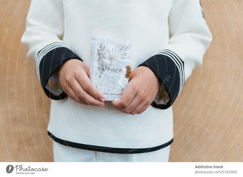 Anonymous child in a white communion suit gently holds a prayer card with religious symbols, expressing the sacredness of First Communion first holy sacrament