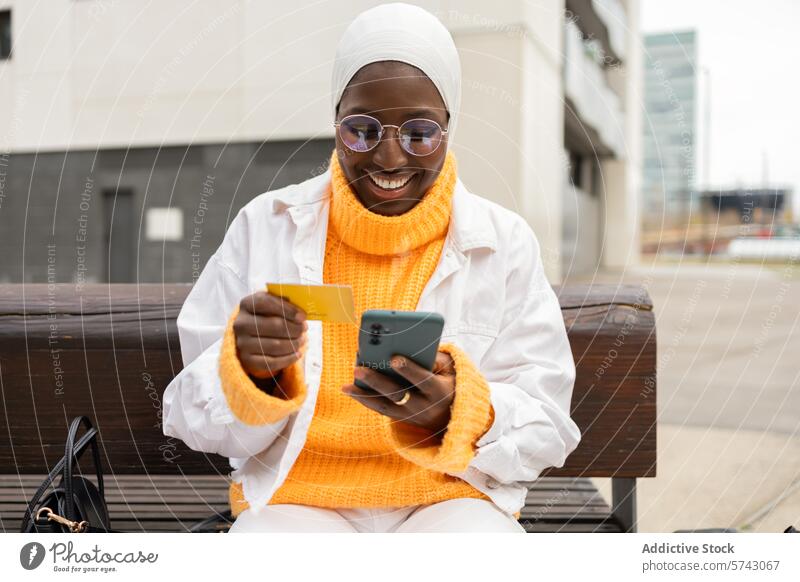 Smiling woman shopping online with card and phone female black african hijab glasses outdoor sitting bench credit card typing muslim purchase smile city urban