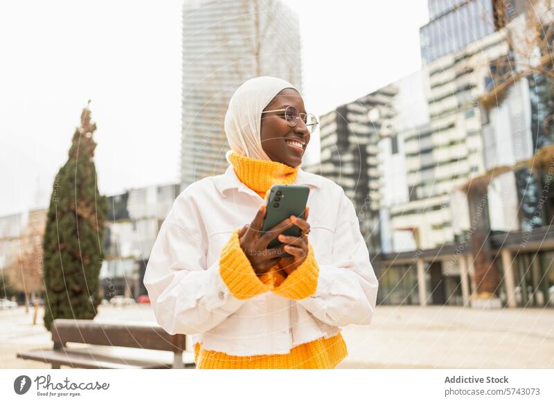 Smiling woman with hijab using phone in urban setting female african smartphone cityscape cheerful headscarf glasses modern building technology communication