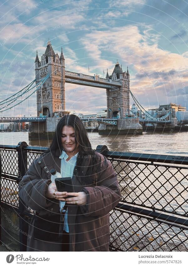 A girl standing in front of Tower Bridge in London, looking at her phone and smiling. tower bridge smile tourist travel travelling london landscape portrait