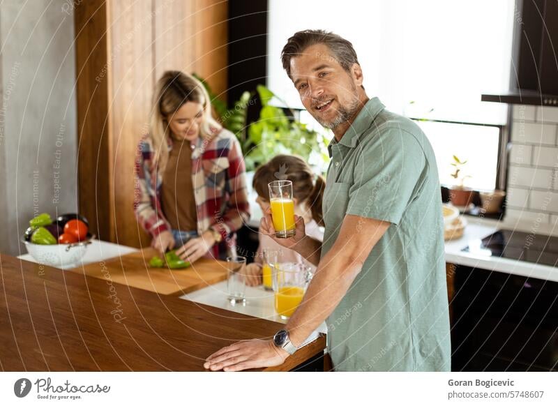Joyful man with a glass of orange juice while a woman and child prepare food in a cozy kitchen family cooking smiling meal preparation domestic life