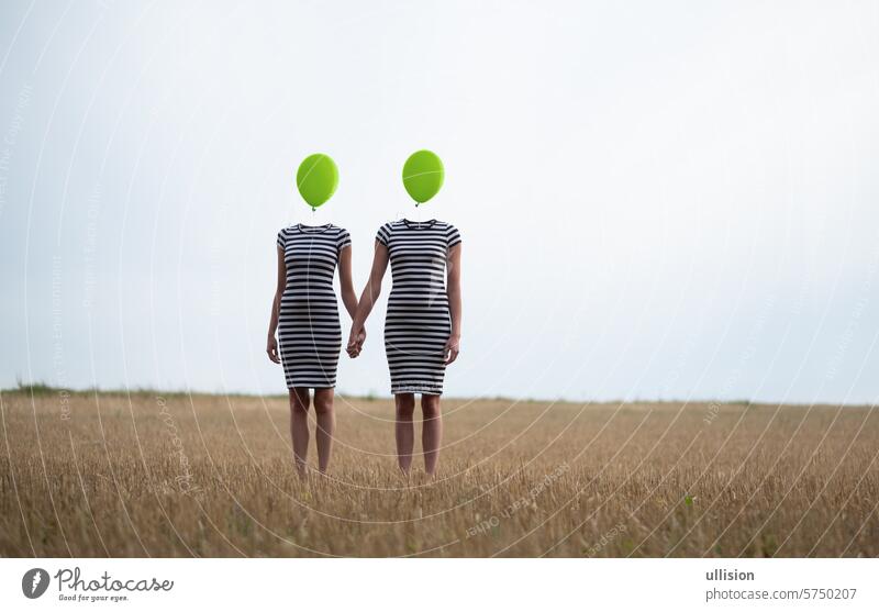 two happy sexy young women, in black and white striped dress, friends, holding hands, heads replaced by green balloons stand hand in hand on harvested corn field