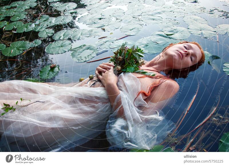 Young sexy seductive crazy longing woman lady redhead Ophelia with curly red hair lies with water lilies, eyes closed in white dress dying in the water in the lake, in lily pond
