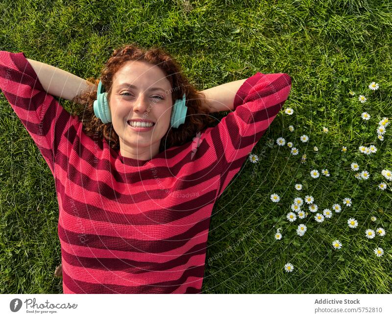 Young redhead woman relaxing on grass with headphones daisies cheerful enjoyment music wireless lying down smiling happiness leisure outdoors nature spring
