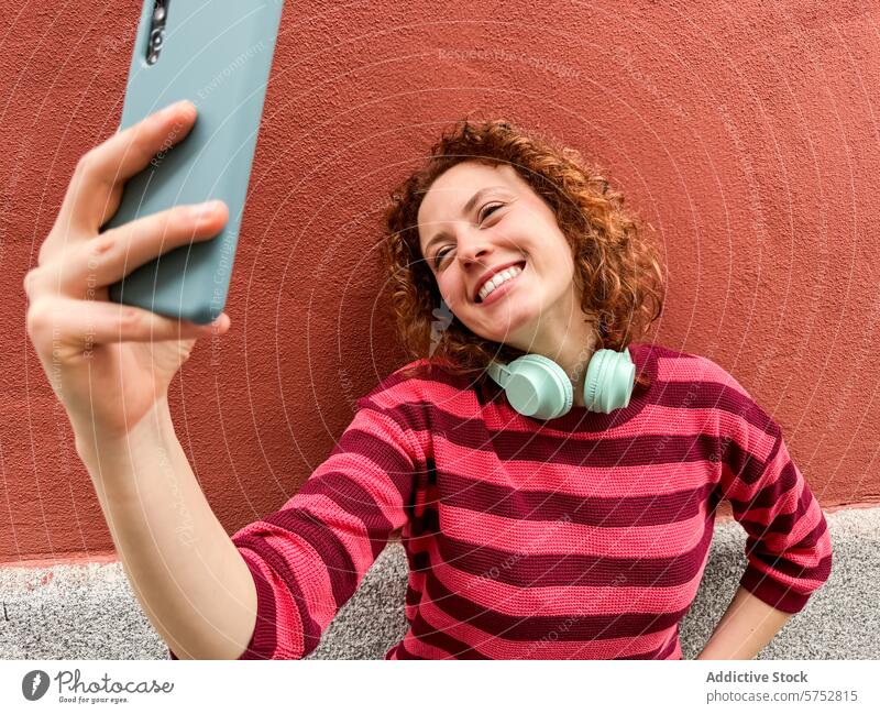 Cheerful redhead taking a selfie with smartphone woman headphones happy cheerful technology social media red wall youth lifestyle casual fashion striped shirt