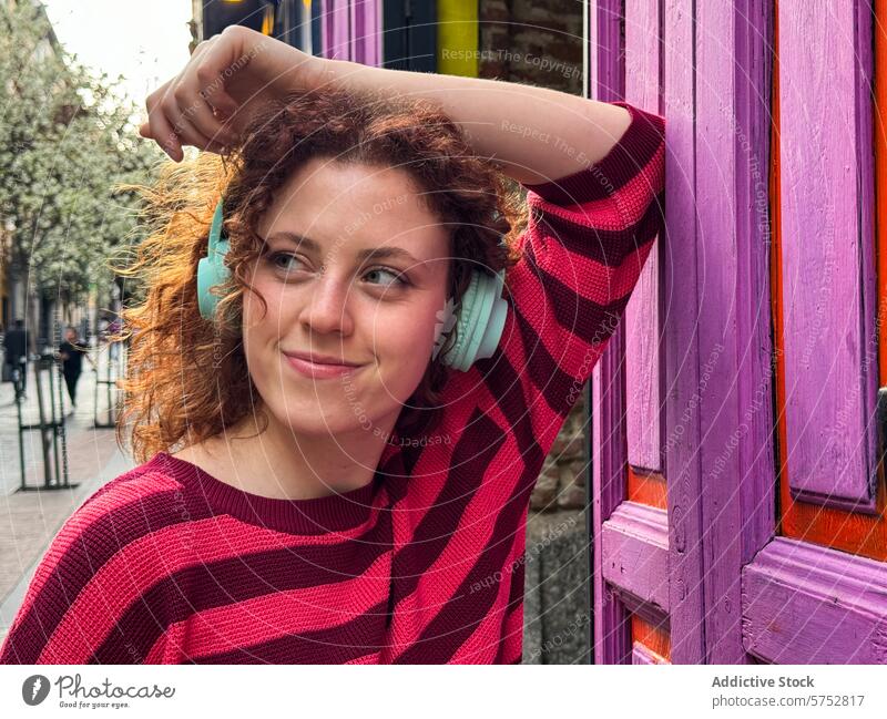 Smiling young redhead woman with headphones leaning on a purple door music smile curly hair striped shirt vibrant city street leisure casual style young adult