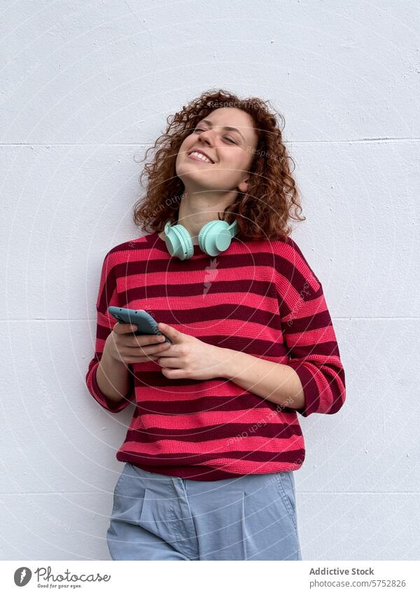 Smiling young redhead woman enjoying music with headphones curly hair smile smartphone white background enjoyment leisure happiness striped sweater fashion