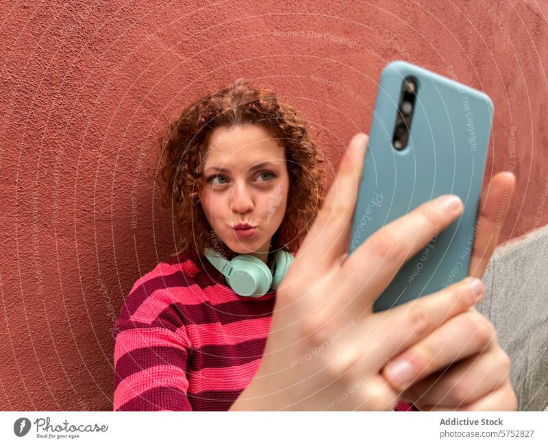 Young redhead woman taking a selfie with a playful expression smartphone curly hair red wall pucker lips holding casual youth photography mobile phone