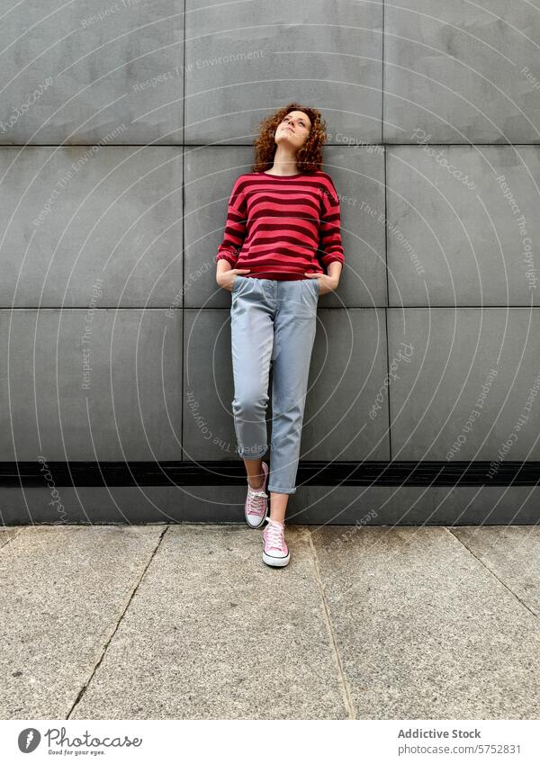 Young redhead woman leaning against a grey wall in casual attire curly hair striped sweater grey pants textured thoughtful fashion style urban streetwear youth