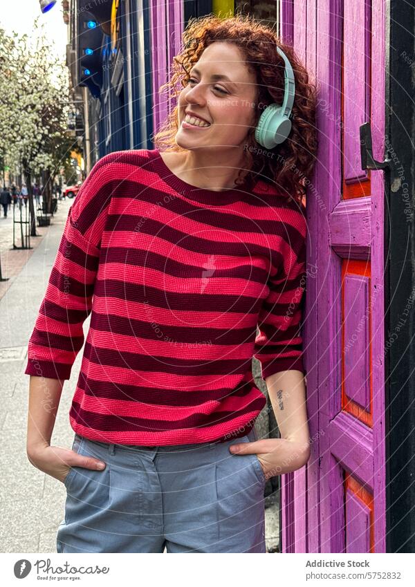 Smiling young redhead woman enjoying music in urban setting headphones smiling door colorful cheerful curly hair striped sweater leaning enjoyment city life