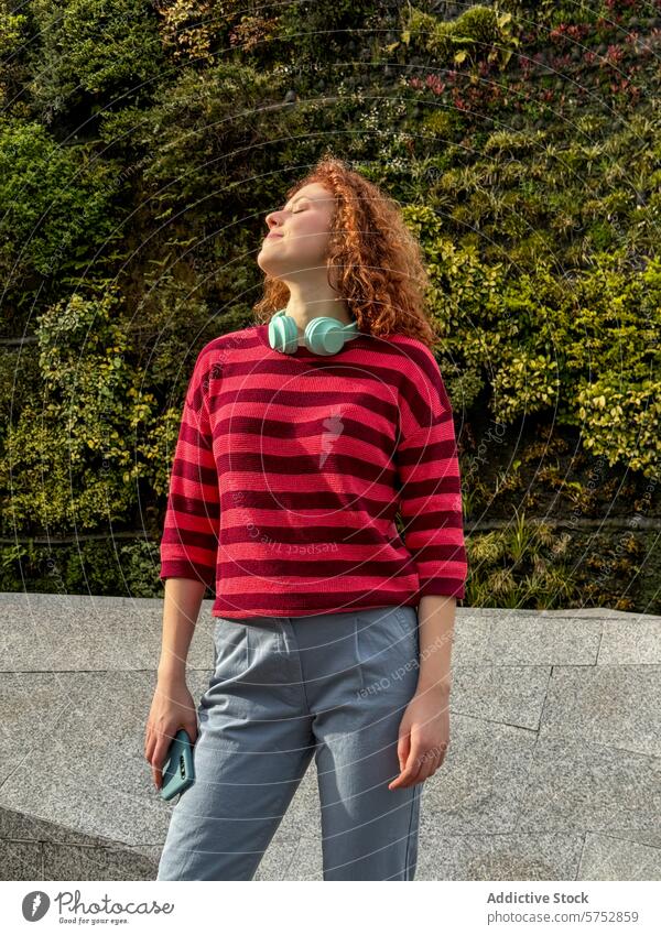 Young redhead woman enjoying the sunshine in casual attire young curly hair smile sunlight headphones smartphone leisure relaxation peaceful enjoyment nature