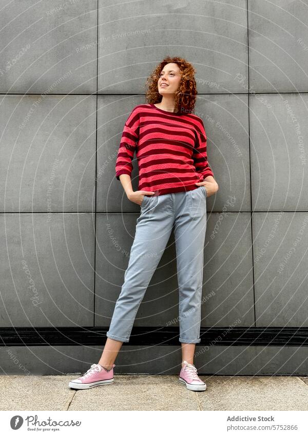 Casual redhead woman in striped sweater posing against wall curly hair smile casual pose gray concrete pant fashion style urban relax cheerful stand outdoor
