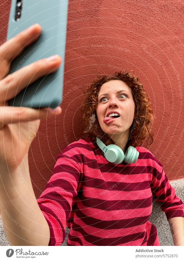 Redhead woman making a funny face while taking a selfie playful grimace smartphone red wall curly hair headphone expression humor casual technology